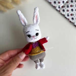 May include: Crocheted white rabbit doll wearing a red jacket, yellow vest, gray pants, and white shoes. The rabbit has small black glasses and a purple bow tie. The doll is handmade.