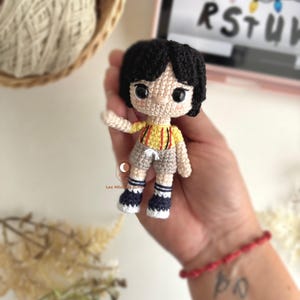 以下が含まれることがあります： 黒い髪のキャラクターの手作りのかぎ針編み人形。赤いストライプの黄色いシャツ、グレーのショーツ、青と白の靴を履いています。人形は手で持たれています。背景には糸のバスケットがあります。