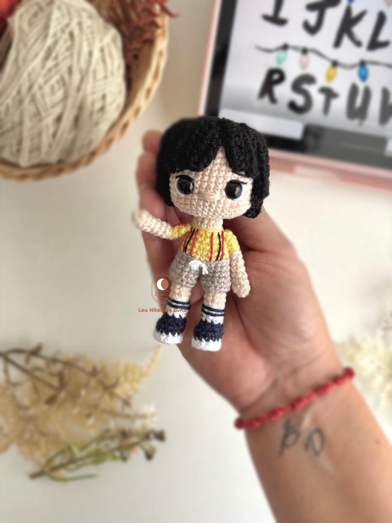以下が含まれることがあります： 黒い髪のキャラクターの手作りのかぎ針編み人形。赤いストライプの黄色いシャツ、グレーのショーツ、青と白の靴を履いています。人形は手で持たれています。背景には糸のバスケットがあります。