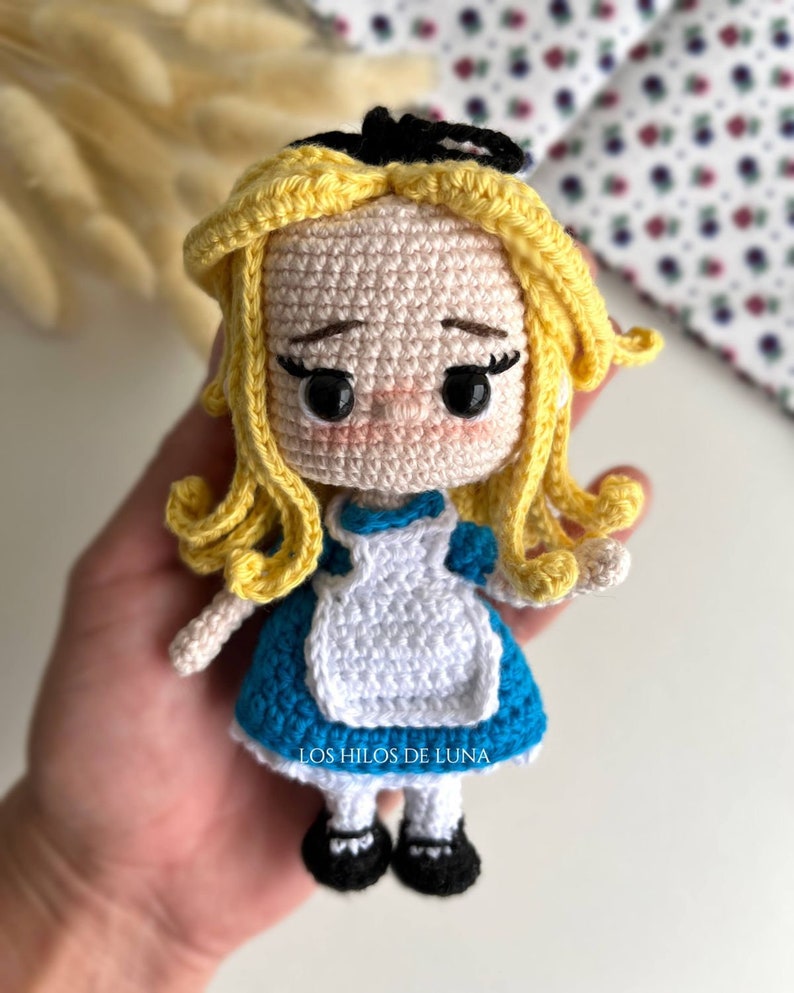 Puede incluir: Mu&ntilde;eca de ganchillo hecha a mano de Alicia en el pa&iacute;s de las maravillas, con un vestido azul, un delantal blanco y cabello rubio. La mu&ntilde;eca tiene zapatos negros y un lazo negro. El texto "LOS HILOS DE LUNA" es visible.