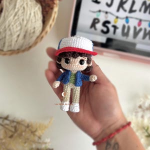 以下が含まれることがあります： 青いジャケット、緑のシャツ、ベージュのズボン、白い靴を履いたキャラクターの手作りのかぎ針編み人形。人形は白と赤の帽子と茶色の髪をしています。人形は手で持たれています。背景には文字とライトが表示されたタブレットが含まれています。