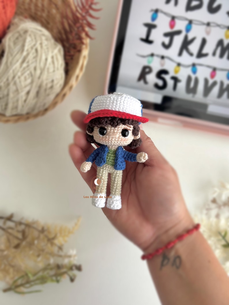 以下が含まれることがあります： 青いジャケット、緑のシャツ、ベージュのズボン、白い靴を履いたキャラクターの手作りのかぎ針編み人形。人形は白と赤の帽子と茶色の髪をしています。人形は手で持たれています。背景には文字とライトが表示されたタブレットが含まれています。