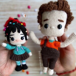May include: Two crocheted amigurumi dolls, one with brown hair and brown overalls and the other with black hair and a teal dress. The dolls are holding their arms out and have big eyes.