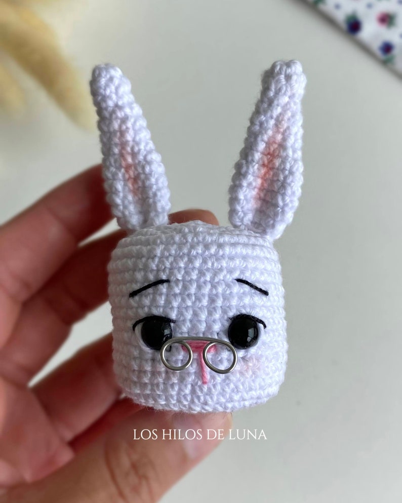 May include: A handmade white crochet bunny head with black eyes, a pink nose, and wire-rimmed glasses. The bunny has long ears with pink accents. The text "LOS HILOS DE LUNA" is at the bottom.