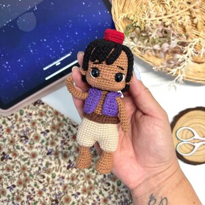 May include: A small, handcrafted crochet doll featuring a tan complexion, black hair, and a red fez hat. The doll wears a purple vest over a brown belt and light beige pants. It has large, dark, glossy eyes and is constructed with visible yarn stitches.