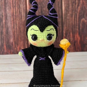 Pode incluir: Uma boneca amigurumi de croch&ecirc; de Mal&eacute;vola de A Bela Adormecida da Disney. A boneca est&aacute; usando um vestido preto com detalhes roxos e tem pele verde, chifres roxos e uma bengala amarela.