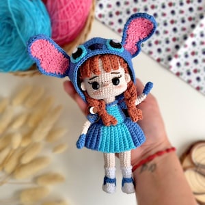 May include: A handmade crochet doll wearing a blue and pink Stitch-inspired hat and dress. The doll has red hair, white socks, and blue shoes. The doll is approximately 15 cm tall.