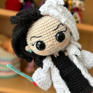 Puede incluir: Mu&ntilde;eca de crochet con pelo blanco y negro, vestida con un abrigo blanco sobre un vestido negro y zapatos rojos. La mu&ntilde;eca tiene grandes ojos negros y una expresi&oacute;n traviesa.