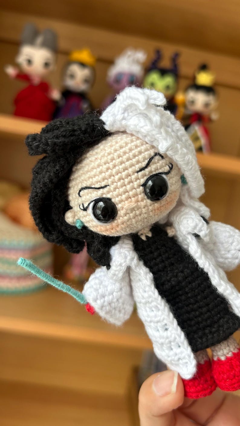 Puede incluir: Mu&ntilde;eca de crochet con pelo blanco y negro, vestida con un abrigo blanco sobre un vestido negro y zapatos rojos. La mu&ntilde;eca tiene grandes ojos negros y una expresi&oacute;n traviesa.