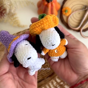 Könnte beinhalten: Zwei handgefertigte Snoopy-Figuren aus Häkelarbeit, eine mit lila Hexenhut und die andere in einem orangefarbenen Kürbiskostüm. Die Figuren sind weiß mit schwarzen Akzenten. Im Hintergrund ein kleiner oranger Kürbis und eine Holzscheibe.