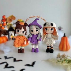 May include: Three handmade crochet dolls dressed in Halloween costumes. The dolls include a pumpkin, a witch, and a ghost. The pumpkin doll wears an orange dress and has a pumpkin on its head. The witch doll wears a purple dress and hat. The ghost doll wears a white outfit.