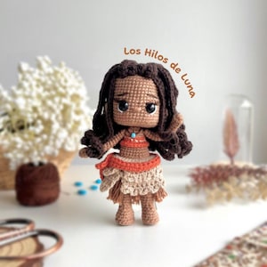 Może przedstawiać: Szydełkowa lalka z ciemnobrązowymi włosami i opaloną cerą. Lalka ma na sobie brązowo-pomarańczowy top i beżową spódnicę z frędzlami. Tekst "Los Hilos de Luna" jest napisany brązowym pismem. Lalka stoi na białej powierzchni.