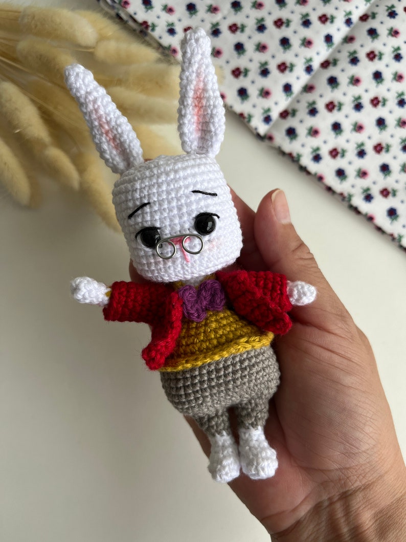 May include: A handmade white crochet rabbit doll with long ears, wearing a red jacket, yellow vest, gray pants, and a purple bow tie. The rabbit has black eyes, a small silver nose ring, and tiny wire-rimmed glasses. The doll is held in a hand.