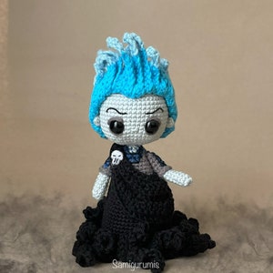 Può includere: Un giocattolo di peluche all'uncinetto di Ade, tratto dal film Disney Hercules. Il giocattolo indossa una tunica nera con un dettaglio di teschio e ossa incrociate. Il giocattolo ha i capelli blu e un viso bianco con occhi neri.