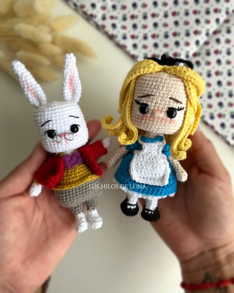 Puede incluir: Dos mu&ntilde;ecos de ganchillo: un conejo blanco con una chaqueta roja y una ni&ntilde;a rubia con un vestido azul. El conejo lleva gafas y una pajarita morada. La ni&ntilde;a tiene un lazo negro en el pelo. Los mu&ntilde;ecos est&aacute;n hechos a mano.