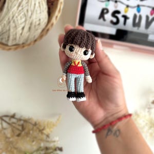 以下が含まれることがあります： 茶色の髪、赤とグレーのシャツ、水色のズボンを着た男の子の手作りのかぎ針編み人形。人形は黒い靴を履いており、手に持っています。背景には、文字I、J、K、L、R、S、T、U、Vが表示されたタブレットが含まれています。