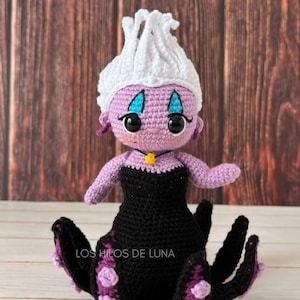 Pode incluir: Uma boneca Ursula de croch&ecirc; da A Pequena Sereia. Ela tem um corpo roxo, cabelo branco, vestido preto e tent&aacute;culos roxos. A boneca est&aacute; sentada em uma superf&iacute;cie de madeira.