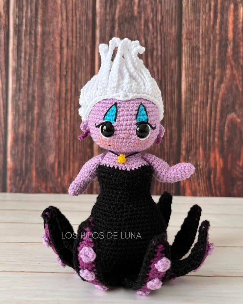 Pode incluir: Uma boneca Ursula de croch&ecirc; da A Pequena Sereia. Ela tem um corpo roxo, cabelo branco, vestido preto e tent&aacute;culos roxos. A boneca est&aacute; sentada em uma superf&iacute;cie de madeira.