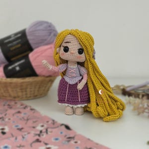 May include: A crocheted doll with long yellow yarn hair, wearing a purple dress with a light purple bodice and pink lace-up details. The doll has large black eyes, a small nose, and pink blush on its cheeks. It features puffed sleeves and a white lace trim at the hem of the skirt, standing upright with one arm raised.