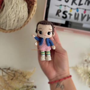 以下が含まれることがあります： ピンクのドレスと青いジャケットを着たキャラクターの手作りのかぎ針編み人形。人形は黒髪で、片手を上げています。人形の高さは約10cmです。背景には、アルファベットとライトが表示されたタブレットが含まれています。