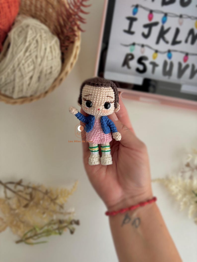 以下が含まれることがあります： ピンクのドレスと青いジャケットを着たキャラクターの手作りのかぎ針編み人形。人形は黒髪で、片手を上げています。人形の高さは約10cmです。背景には、アルファベットとライトが表示されたタブレットが含まれています。