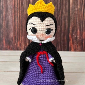 Pode incluir: Boneca de croch&ecirc; da Rainha M&aacute; com uma coroa amarela, um vestido roxo, uma capa preta e uma fita vermelha. A boneca tem um colar branco e um rosto rosa com olhos pretos.