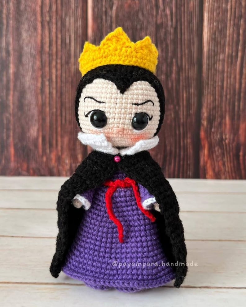 Pode incluir: Boneca de croch&ecirc; da Rainha M&aacute; com uma coroa amarela, um vestido roxo, uma capa preta e uma fita vermelha. A boneca tem um colar branco e um rosto rosa com olhos pretos.