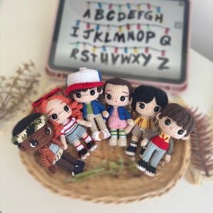 Puede incluir: Una colección de muñecos de ganchillo hechos a mano, cada uno representando personajes de una popular serie de ciencia ficción. Los muñecos están dispuestos en una cesta tejida, con una tableta que muestra el alfabeto en el fondo. Los muñecos visten trajes a juego.