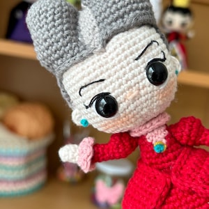 May include: A crocheted doll with gray hair, a white face, and large black eyes. The doll is wearing a red dress with a pink collar and a gold brooch. The doll is holding out its right hand.