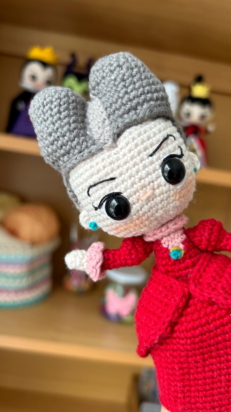 May include: A crocheted doll with gray hair, a white face, and large black eyes. The doll is wearing a red dress with a pink collar and a gold brooch. The doll is holding out its right hand.