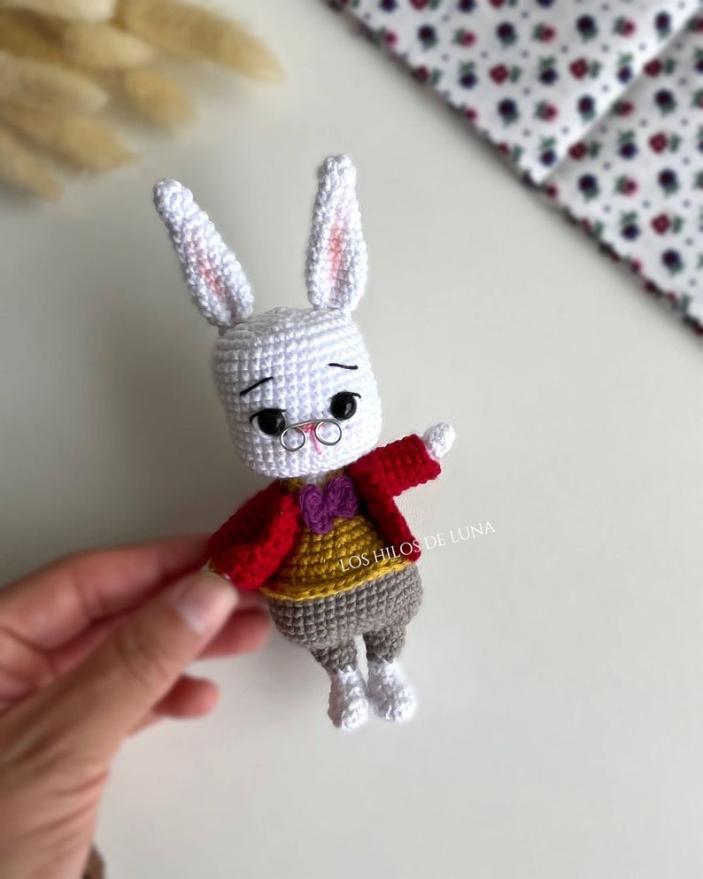 Puede incluir: Mu&ntilde;eco de conejo blanco tejido a ganchillo, con chaqueta roja, chaleco amarillo, pantal&oacute;n gris y zapatos blancos. El conejo tiene gafas redondas y una pajarita morada. Hecho a mano.