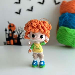 Pode incluir: Uma boneca de crochê com cabelo encaracolado laranja, vestindo uma camisa amarela e verde, calças marrons e sapatos azuis. A boneca está em pé sobre uma superfície branca.