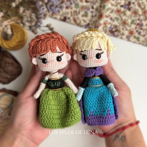 May include: Two crocheted dolls, one with a green dress and the other with a blue dress, are holding hands. The dolls have brown and blonde hair, respectively. The dolls are wearing crowns and have embroidered facial features.