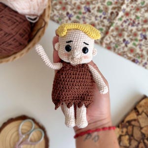 May include: Crocheted doll with brown hair, brown clothing, and a white face. The doll is holding its right arm up in the air.