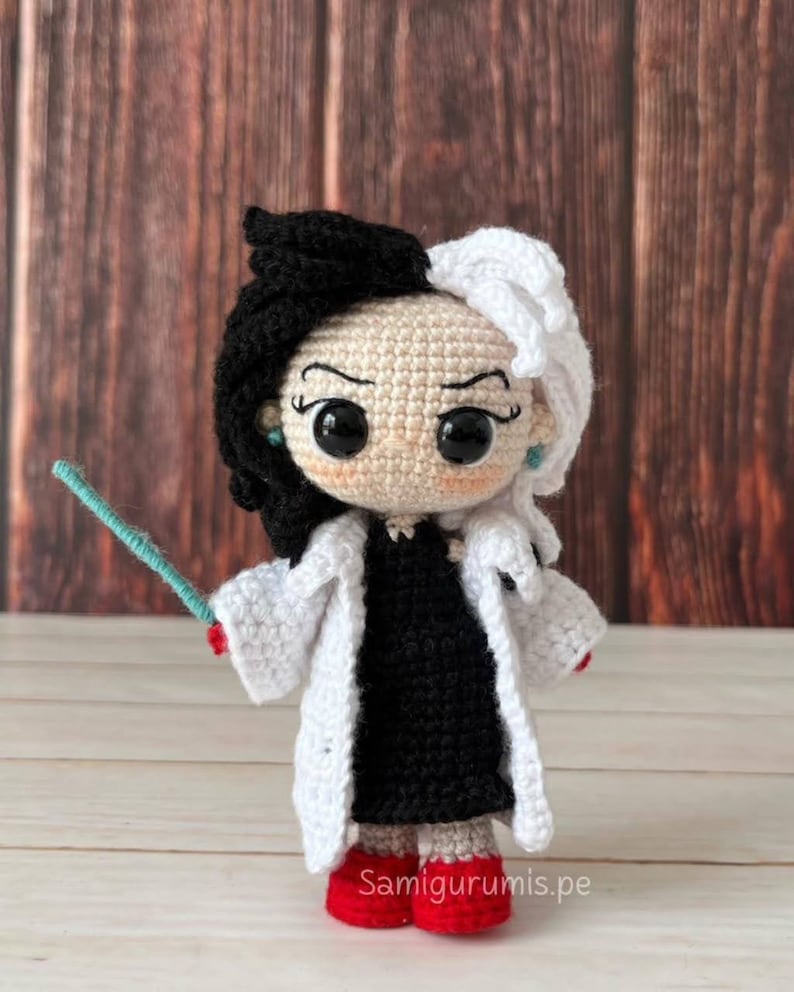 Pode incluir: Boneca de croch&ecirc; da Cruella de Vil do filme 101 D&aacute;lmatas da Disney. A boneca est&aacute; usando um vestido preto, um casaco branco e sapatos vermelhos. Ela tem cabelo preto e branco e est&aacute; segurando uma varinha azul.