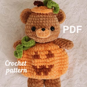 Op de afbeelding: Een bruine gehaakte berenknuffel in een oranje pompoenkostuum met een jack-o'-lantern gezicht, groene ranken en een bruine steel op het hoofd. De tekst PDF en Crochet pattern zijn in het wit op de afbeelding zichtbaar.