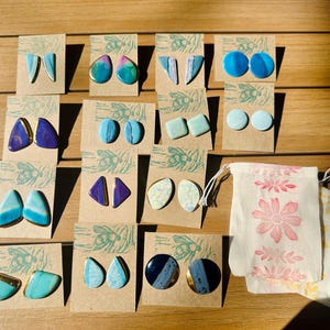 Op de afbeelding: Assortiment van handgemaakte keramische oorbellen in verschillende tinten blauw en paars. De oorbellen worden gepresenteerd op kleine kaartjes met een bijenontwerp. De oorbellen worden verpakt in een witte stoffen tas met een bloemenpatroon.