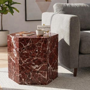 May include: A hexagonal side table with a red marble design. A small notebook, a cup of coffee, and a decorative item are on top. The table is next to a gray sofa and a potted plant.