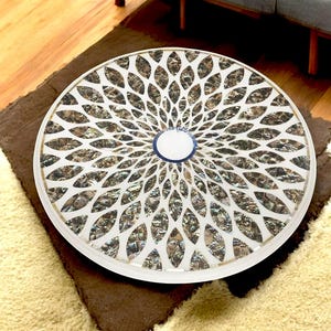 Puede incluir: Una mesa redonda de madera blanca con un patrón decorativo de hojas de nácar superpuestas e iridiscentes en tonos marrones, dorados y verdes. El centro de la mesa tiene un círculo azul y blanco.