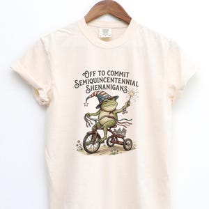 Könnte beinhalten: Cremefarbenes T-Shirt mit einer skurrilen Illustration einer Frosch auf einem Dreirad. Der Frosch trägt einen patriotischen Hut und hält einen Wunderkerze. Der Text auf dem Shirt lautet: "Off to Commit Semiquincentennial Shenanigans."
