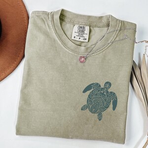 May include: A sage green Comfort Colors t-shirt with a teal turtle graphic. A silver necklace with a pink pendant rests on the shirt. A brown hat and dried palm leaves are in the background.