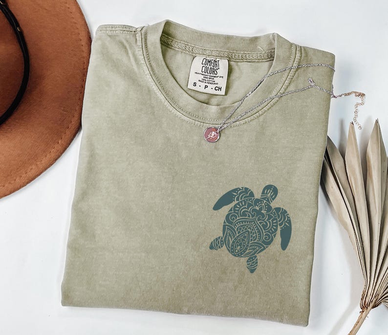 May include: A sage green Comfort Colors t-shirt with a teal turtle graphic. A silver necklace with a pink pendant rests on the shirt. A brown hat and dried palm leaves are in the background.