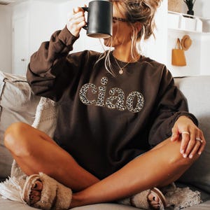 May include: A brown sweatshirt with the word "ciao" in a leopard print design. The person is holding a black mug. The person is wearing fuzzy slippers and sitting on a couch.