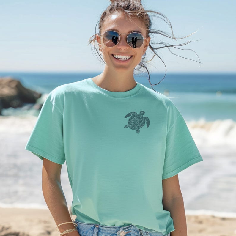 May include: A light teal t-shirt featuring a small, dark gray turtle design on the upper left chest. The shirt has short sleeves and a relaxed fit. The background shows a beach scene with the ocean and blue sky.