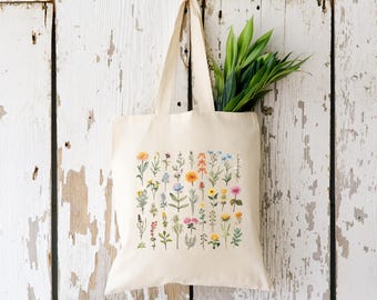 May include: Cream-colored tote bag with a colorful floral print. The design features various wildflowers in shades of yellow, orange, pink, and blue. The bag is hanging against a white wooden background, and has green leaves inside.
