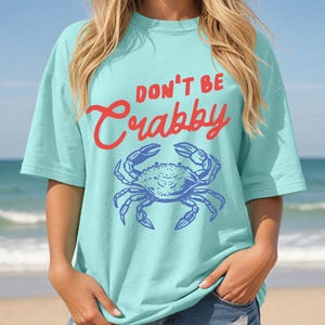 May include: A light blue t-shirt with the words "DON'T BE Crabby" in red script above a blue crab illustration. The shirt has short sleeves and a relaxed fit, perfect for a day at the beach.