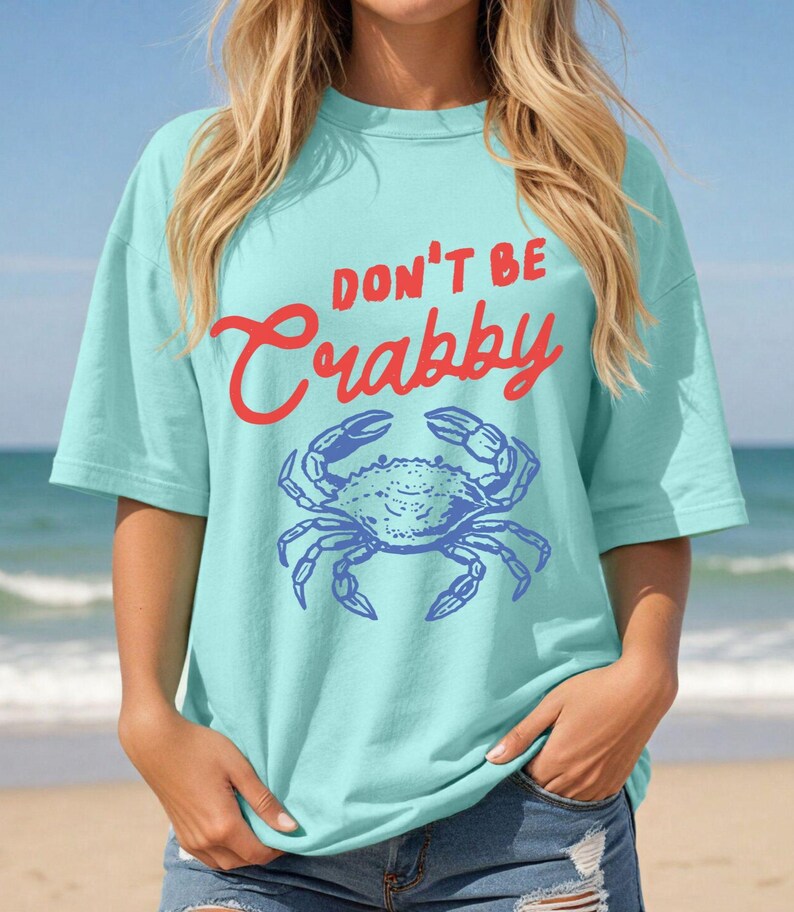 May include: A light blue t-shirt with the words "DON'T BE Crabby" in red script above a blue crab illustration. The shirt has short sleeves and a relaxed fit, perfect for a day at the beach.