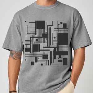 May include: Gray t-shirt featuring a black geometric design of squares, rectangles, lines, and circles. The short-sleeved shirt has a relaxed fit and a crew neck. The design is centered on the front of the shirt.