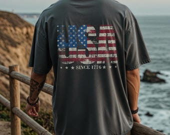 May include: Gray t-shirt with a distressed USA flag design on the back. The flag features red and white stripes and blue stars. The text "SINCE 1776" is below the flag. The shirt is a relaxed fit.