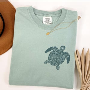 May include: A light green Comfort Colors t-shirt featuring a detailed teal turtle design. A gold necklace with a pendant rests on the shirt. A brown hat and dried palm leaves are in the background.